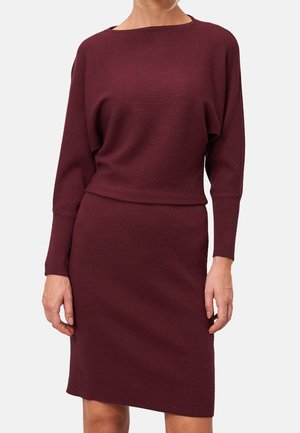 Woman wearing a ribbed, long-sleeve, burgundy knit dress with a fitted waist and knee-length skirt on a light background.