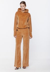 Zestaw welurowej bluzy z kapturem i spodni szerokich w ciepłym odcieniu brązu, z przednim złotym logo. Gładka tekstura, luźny krój i długie rękawy.