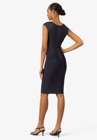 Navyblaues, enganliegendes Kleid mit horizontaler Rippstruktur, kurzen Ärmeln und verdecktem Reißverschluss auf der Rückseite. Kombiniert mit schwarzen Absatzsandalen.