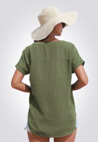 Camisa de manga corta texturizada en verde oliva con detalle en la costura trasera, combinada con pantalones cortos de mezclilla. Se lleva un sombrero de paja de ala ancha.