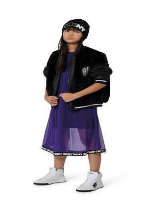 Zwarte nep bont bomberjack, paarse doorschijnende jurk met DKNY-logo aan de zoom, witte high-top sneakers en zwarte beanie met witte letters.