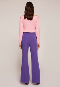 Kvinna med vågigt brunt hår som bär en rosa off-shoulder tröja och högmidjade lila vida byxor, stående mot en enfärgad bakgrund.