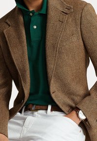 Hnědý blazer v herringbonovém vzoru se strukturovanými klopami, nošený přes tmavě zelené polo. Bílé kalhoty a hnědý kožený opasek se zlatou přezkou. Viditelný stříbrný hodinky.