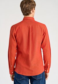 Koszula z długim rękawem w jednolitym spalonym pomarańczowym kolorze, wykonana z teksturowanej tkaniny. Posiada klasyczny kołnierzyk oraz mankiety zapinane na guziki na zwężających się rękawach.