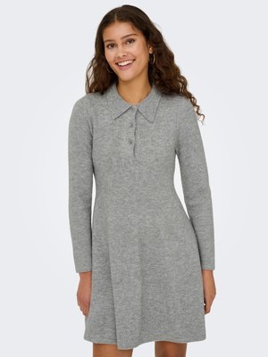 Femme souriante aux longs cheveux bouclés porte une robe en maille grise à manches longues avec un col et trois boutons.
