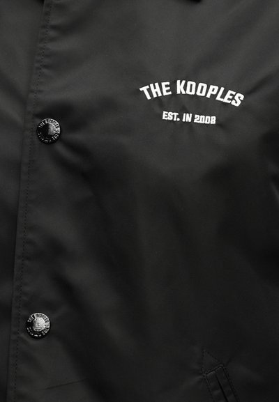 Μαύρο μπουφάν με λευκό κεντημένο κείμενο «THE KOOPLES EST. IN 2008» και μαύρα κουμπιά με ανάγλυφο το όνομα της μάρκας.