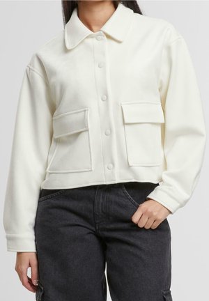 Femme portant une veste crème boutonnée avec deux grandes poches avant, associée à un jean noir taille haute.