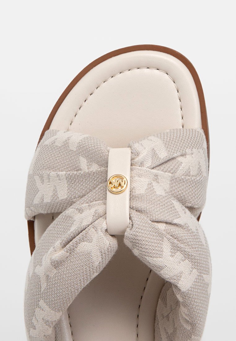 Sandale beige clair avec une sangle en tissu texturé, ornée d'un nœud et d'un accent logo. La semelle est lisse et marron.