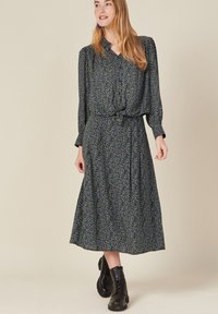 Schwarzes florales Midikleid mit langen Ärmeln, Bindegürtel und auffälligem lila-grünen Muster. Tragen mit schwarzen Knöchelstiefeln.