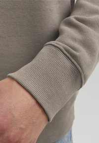 Manica di felpa marrone chiaro con polsino a costine testurizzate, realizzata in tessuto morbido e con un design aderente. Dettaglio della mano in primo piano visibile.