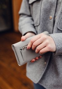 Gray leather wallet held in hands, featuring a zippered pocket and smooth texture. The wallet has clean lines and a compact design.