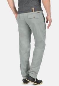 Pantalon gris clair à coupe décontractée, avec deux poches arrière à fermeture par bouton et une étiquette sur la ceinture. Porté avec des chaussures foncées.