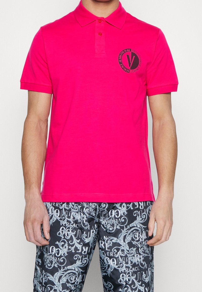 Homme portant un polo rose vif avec un logo circulaire noir sur la poitrine et un pantalon noir à motifs avec des dessins blancs complexes et des inscriptions.