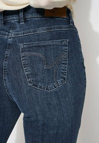 Jeans en denim bleu foncé présentant un motif de couture courbé distinctif sur la poche arrière et une étiquette de marque en cuir sur la taille.