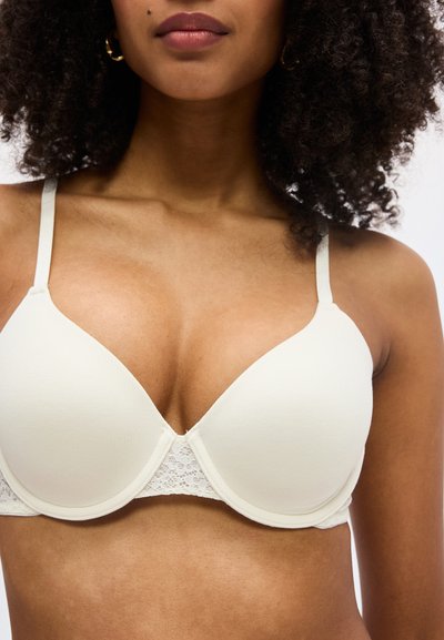 Κοντινό πλάνο γυναίκας που φοράει ένα εκρού πάπλωμα σουτιέν με δαντελένιες λεπτομέρειες και λεπτές ρυθμιζόμενες τιράντες, που δείχνει τον λαιμό και το πάνω μέρος του κορμού της.