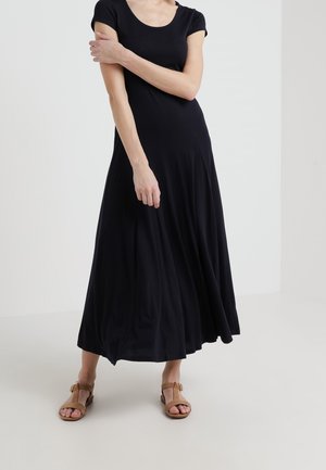 Kobieta stoi, ubrana w czarną maxi sukienkę z krótkim rękawem oraz brązowe, odkryte sandały na jednolitym białym tle.