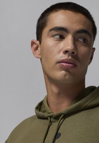 Felpa verde oliva con cappuccio e cordino. Il tessuto è liscio con una texture sottile. Il design presenta un logo nero sulla parte frontale.