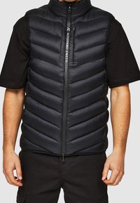 Sort polstret vest med høj krave, vandret quiltet design, lynlås foran og logodetalje på lynlåsen. Bærer den over en sort skjorte.