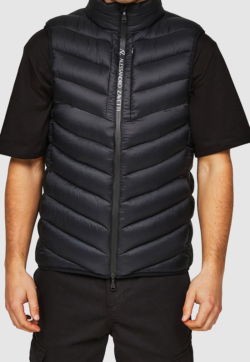 Sort polstret vest med høj krave, vandret quiltet design, lynlås foran og logodetalje på lynlåsen. Bærer den over en sort skjorte.