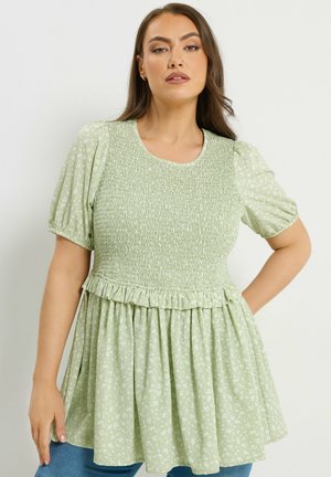 Frau mit langem braunem Haar, die ein hellgrünes, geblümtes, gerafftes Oberteil mit kurzen Puffärmeln und ausgestelltem Saum trägt und vor weißem Hintergrund posiert.