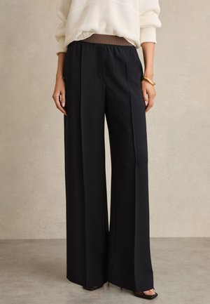 Mujer que lleva pantalones anchos negros con cintura elástica marrón, suéter de punto blanco, pulsera dorada y tacones abiertos negros.