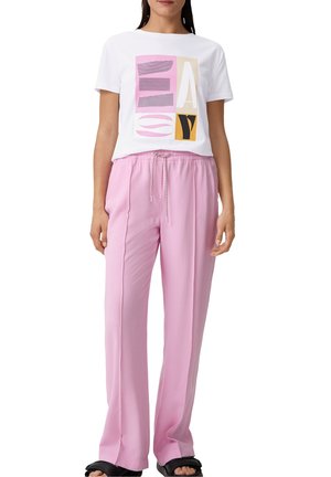 Kobieta w białej koszuli z krótkim rękawem z kolorowym abstrakcyjnym wzorem oraz luźnych różowych spodniach zawiązywanych w pasie, zestawionych z czarnymi sandałami na obcasie.