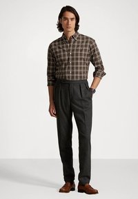 Καφέ καρό πουκάμισο με κουμπιά, γκρι παντελόνια με πιέτες και καφέ δερμάτινα παπούτσια. Το μοντέλο στέκεται με τα χέρια στις τσέπες μπροστά σε ένα απλό φόντο.