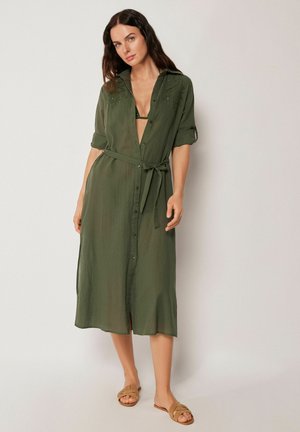Kobieta ubrana w oliwkowozieloną, zapinaną na guziki sukienkę midi z paskiem w talii i podwiniętymi rękawami, w połączeniu z jasnobrązowymi klapkami, stoi na tle białej ściany.
