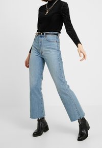 Helle blaue, hoch geschnittene Schlagjeans mit fransigem Saum, kombiniert mit einem schwarzen gerippten Langarmoberteil und schwarzen Stiefeletten.