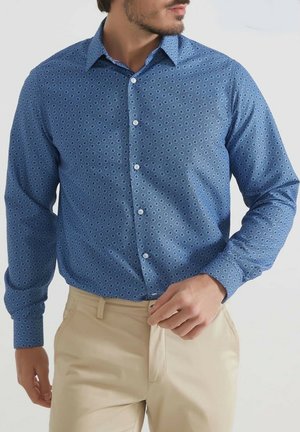 Chemise boutonnée bleue avec une texture à motifs circulaires, manches longues et col classique, associée à un pantalon beige.