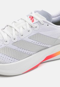 Hvid atletisk sko med en tekstureret mesh-overdel, sølvdetaljer og en polstret sål med en gradient i røde og orange nuancer.