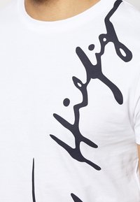 Camiseta de algodón blanca con un atrevido diseño gráfico abstracto en negro en la parte delantera, que enfatiza líneas curvas y grosores variables.