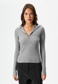 Maglione grigio a maniche lunghe con collo alla pescatora e zip, dalla silhouette aderente. Tessuto morbido con un motivo testurizzato. Abbinato a pantaloni neri.