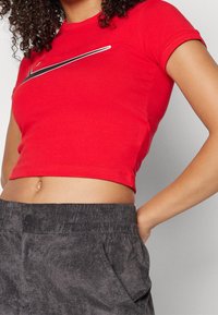 Czerwony, krótki top z krótkim rękawem wykonany z bawełny, z okrągłym dekoltem. Na piersiach znajduje się czarne logo Nike. Połączony z ciemnoszarymi, teksturowanymi spodniami.