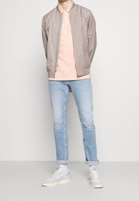 Helle beiges Bomberjacke, hellrosa Polo, hellblaue Jeans, weiße Sneakers mit grauen Akzenten. Klassischer Schnitt, lässiger Stil.