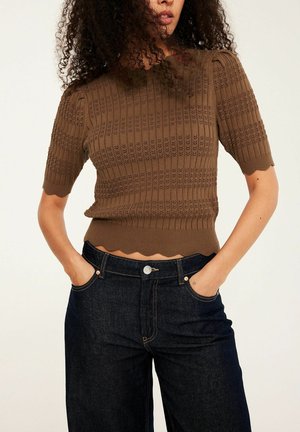 Kobieta o kręconych włosach, ubrana w brązowy krótki sweter z falistym dołem i ciemnoniebieskie, wysokie dżinsy, ręce w kieszeniach.