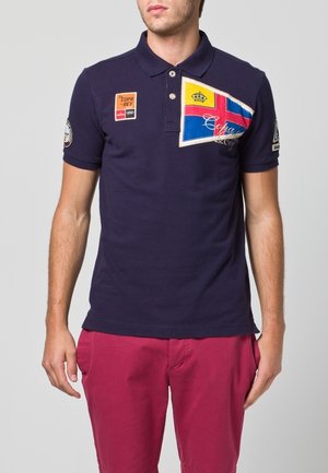 Granatowa koszulka polo z naszywkami, w tym kolorową flagą i logo na rękawach. Krótkie rękawy i klasyczny kołnierzyk.