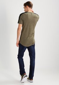Camisa de manga corta verde oliva con acentos negros en los hombros, dobladillo curvo y detalle de logo en la parte posterior, combinada con jeans azul oscuro y zapatillas verdes.