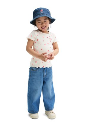 Lächendes Kind mit einem blauen Anglerhut, weißem Hemd mit roten Herzen, lockeren blauen Jeans und beigen Sneakers vor weißem Hintergrund stehend.