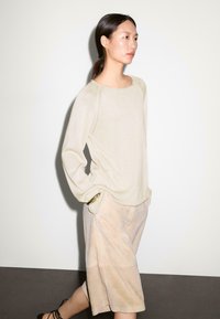 Maglione di maglia beige con collo rotondo e maniche raglan, abbinato a pantaloni corti color beige chiaro. Accentatura in oro notevole sui pantaloni corti.