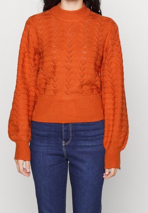 Personne portant un pull en tricot orange avec un motif texturé et des manches longues bouffantes, associé à un jean bleu foncé.