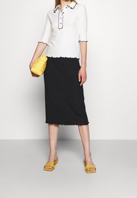 Bílý žebrovaný top s černým lemováním ve tvaru vlny, detaily na knoflících a tříčtvrtečními rukávy, v kombinaci s černou sukní s lemováním ve tvaru vlny. Žlutá pletená taška.