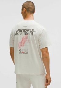 T-shirt blanc à manches courtes avec un tissu texturé, impression au dos avec un texte en gras noir et rouge, au design minimaliste et avec un branding subtil.