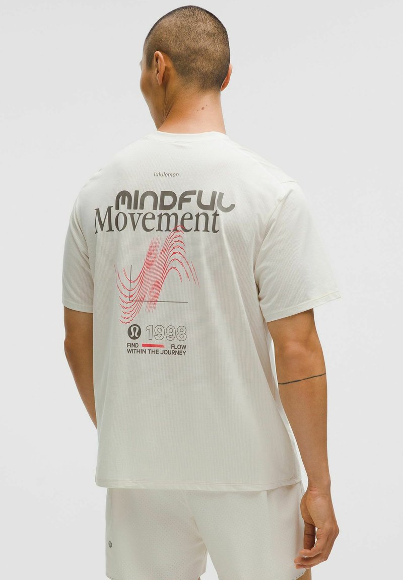 T-shirt blanc à manches courtes avec un tissu texturé, impression au dos avec un texte en gras noir et rouge, au design minimaliste et avec un branding subtil.