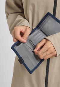 Gray wallet with navy trim, featuring multiple card slots and a zippered compartment, held by a hand wearing a tattoo. Soft fabric texture.