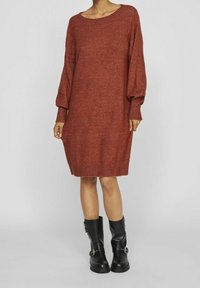 Robe tricotée couleur rouille avec manches longues, coupe ample, et poignets côtelés. Associée à des bottes en cuir noires montant jusqu'aux genoux avec des boucles.