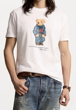 Hombre con camiseta blanca con gráfico de un oso de peluche con ropa vaquera, combinado con vaqueros azules desgastados y cinturón de cuero marrón.
