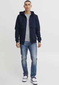 Felpa blu navy con zip e tasche frontali, indossata sopra una maglietta grigia. Abbinata a jeans blu sbiaditi e sneakers bianche con dettagli blu navy.