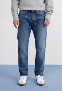 Vaqueros de mezclilla azul con corte de pierna recta, que cuentan con un diseño clásico de cinco bolsillos y un desgaste sutil. Combinados con zapatillas blancas.