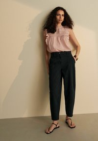 Blouse rose clair à manches volantées, pantalons noirs taille haute et sandales plates noires avec un accent doré. Tissus lisses, coupe ajustée.